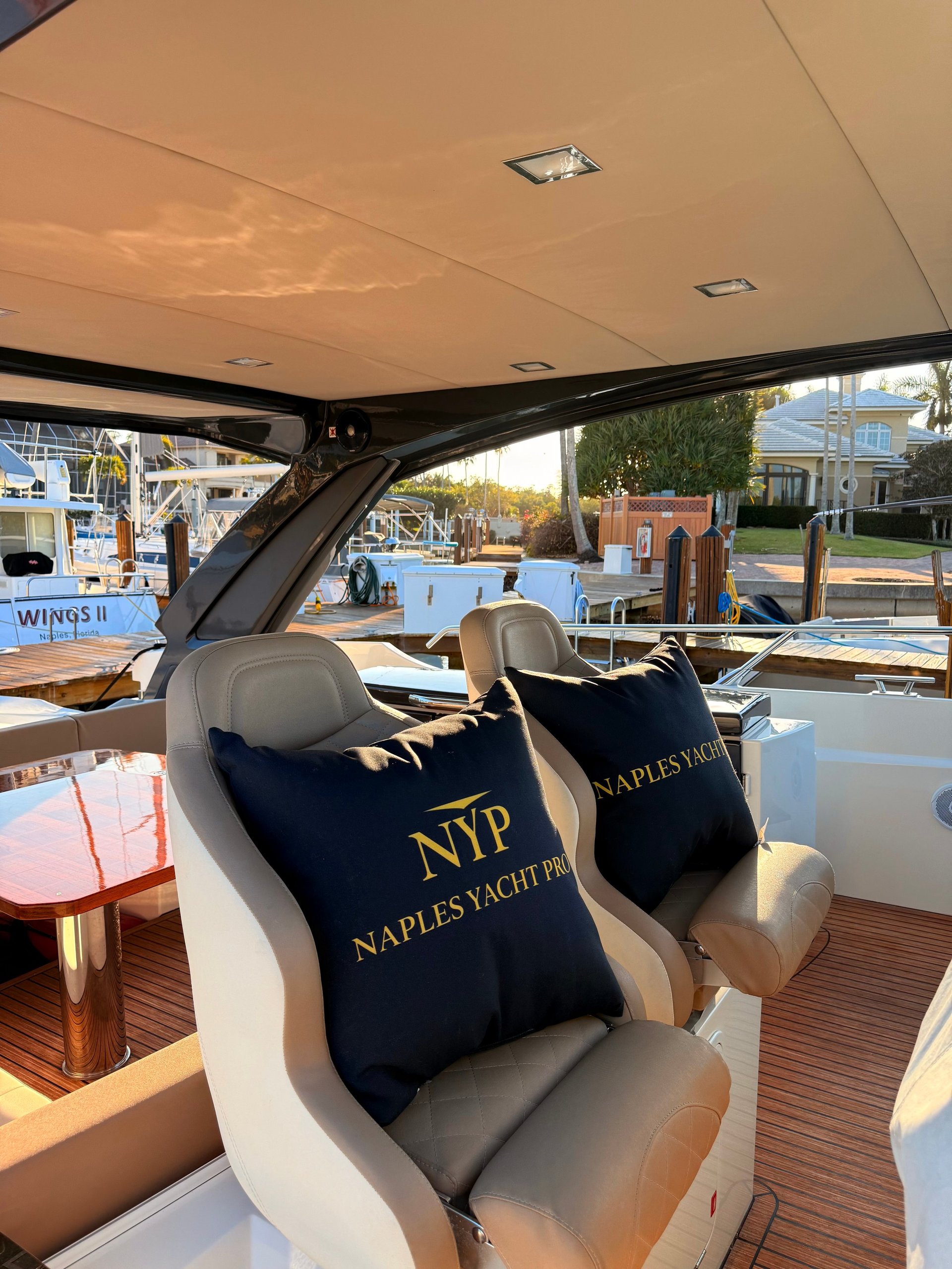 Interior view of a boat cabin with gray seats, black embroidered pillows labeled Naples Yacht, and a view of the marina and docked boats outside